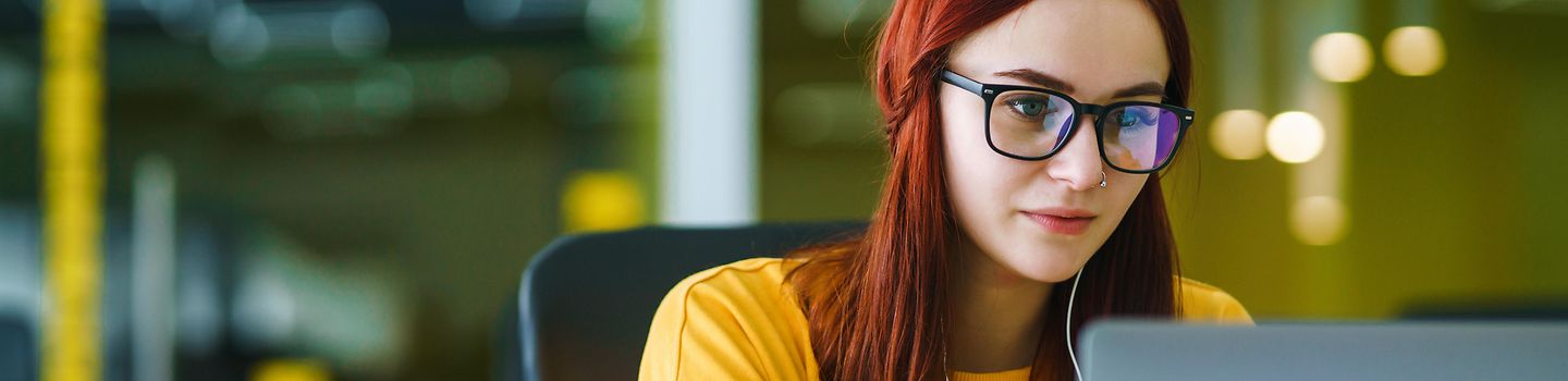 Eine junge Frau mit roten Haare, gelben Pullover und einer Brille sitzt am Tisch und arbeitet an einem Laptop. Links neben ihr auf dem Tisch befinden sich Bücher und Zettel und ein Glas mit Stiften.