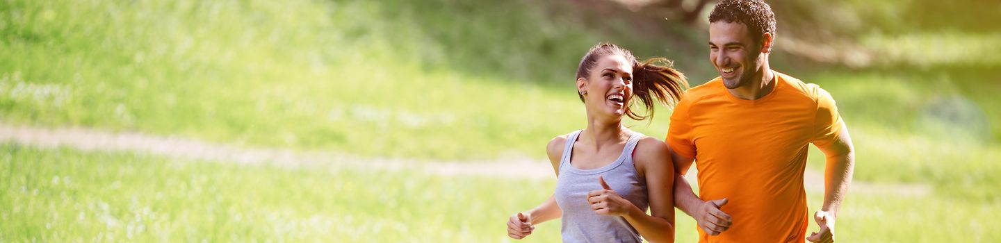 Eine junge Frau und ein junger Mann joggen nebeneinander über einen Weg durch eine Rasenfläche. Sie tragen Sportkleidung und lachen. Sie sieht in seine Richtung. Die Sonne scheint.