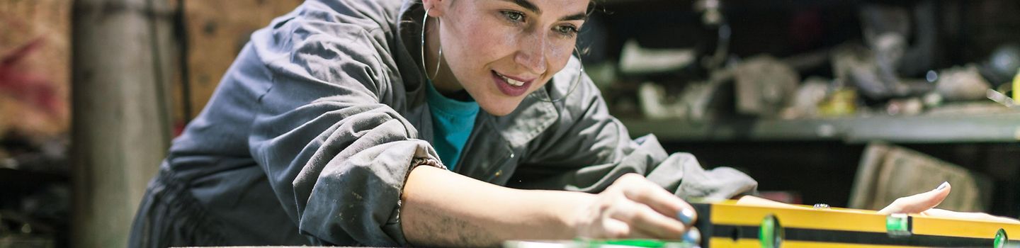 Eine Frau in einer Werkstatt bückt sich über die Werkbank und greift zur Wasserwaage. Eine Frau in einer Werkstatt bückt sich über die Werkbank und greift zur Wasserwaage.