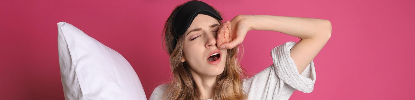 Junge Frau mit Schlafbrille auf dem Kopf und Kopfkissen unter dem Arm gehängt. Über ihr ein Piktogramm, das eine leere Batterie zeigt.