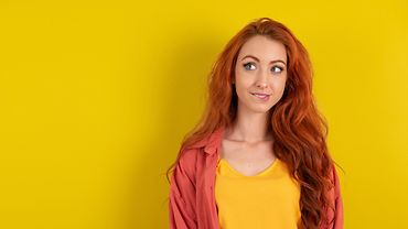 Junge Frau mit langen roten Haaren vor gelbem Hintergrund, sie beißt sich auf die Lippe und schaut vorsichtig optimistisch zur Seite.